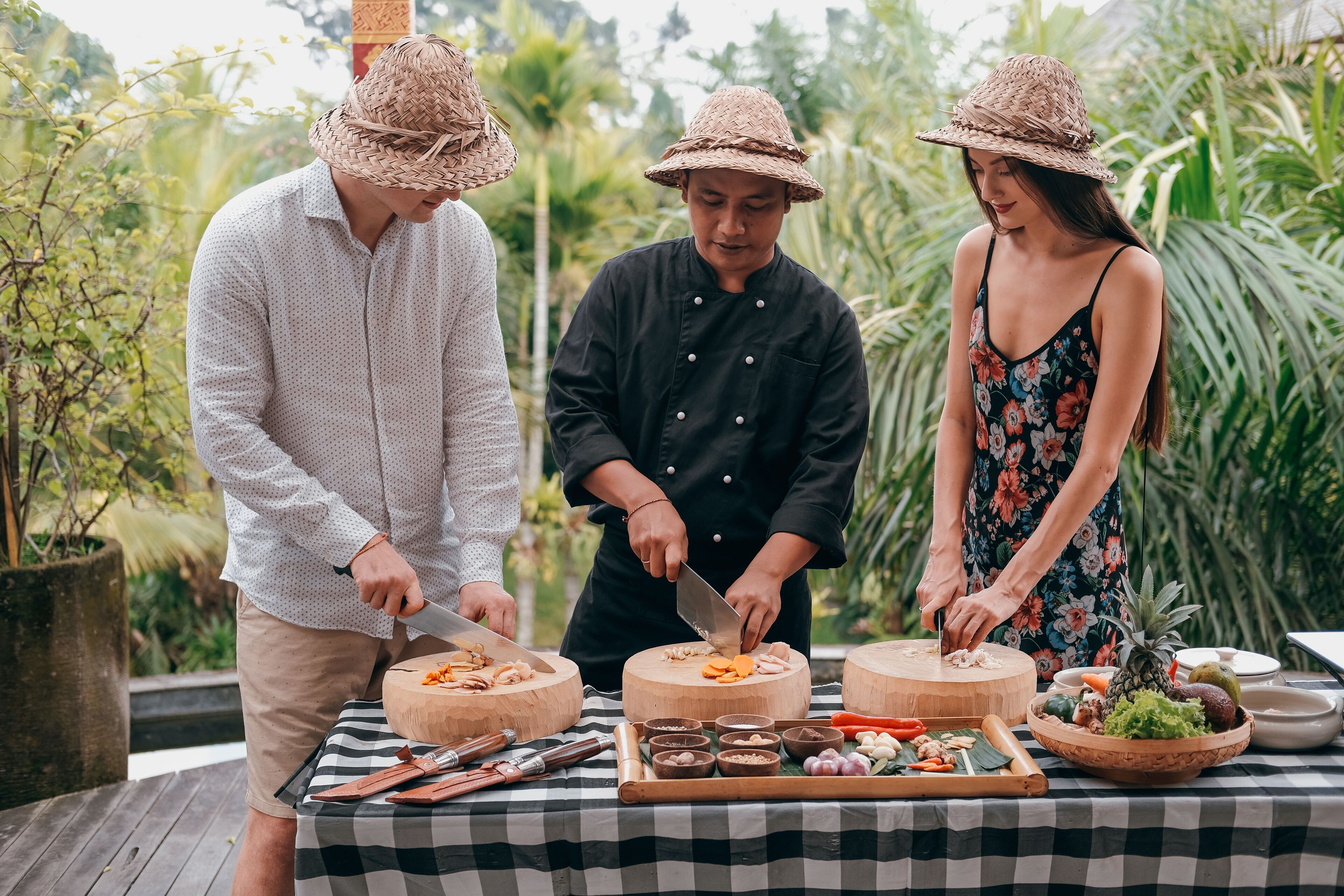 Three people preparing food outdoors with a checkered tablecloth, wooden cutting boards, and various ingredients.