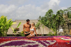 A man and a woman sitting on a flower-covered area with tropical plants and a thatched-roof structure in the background.