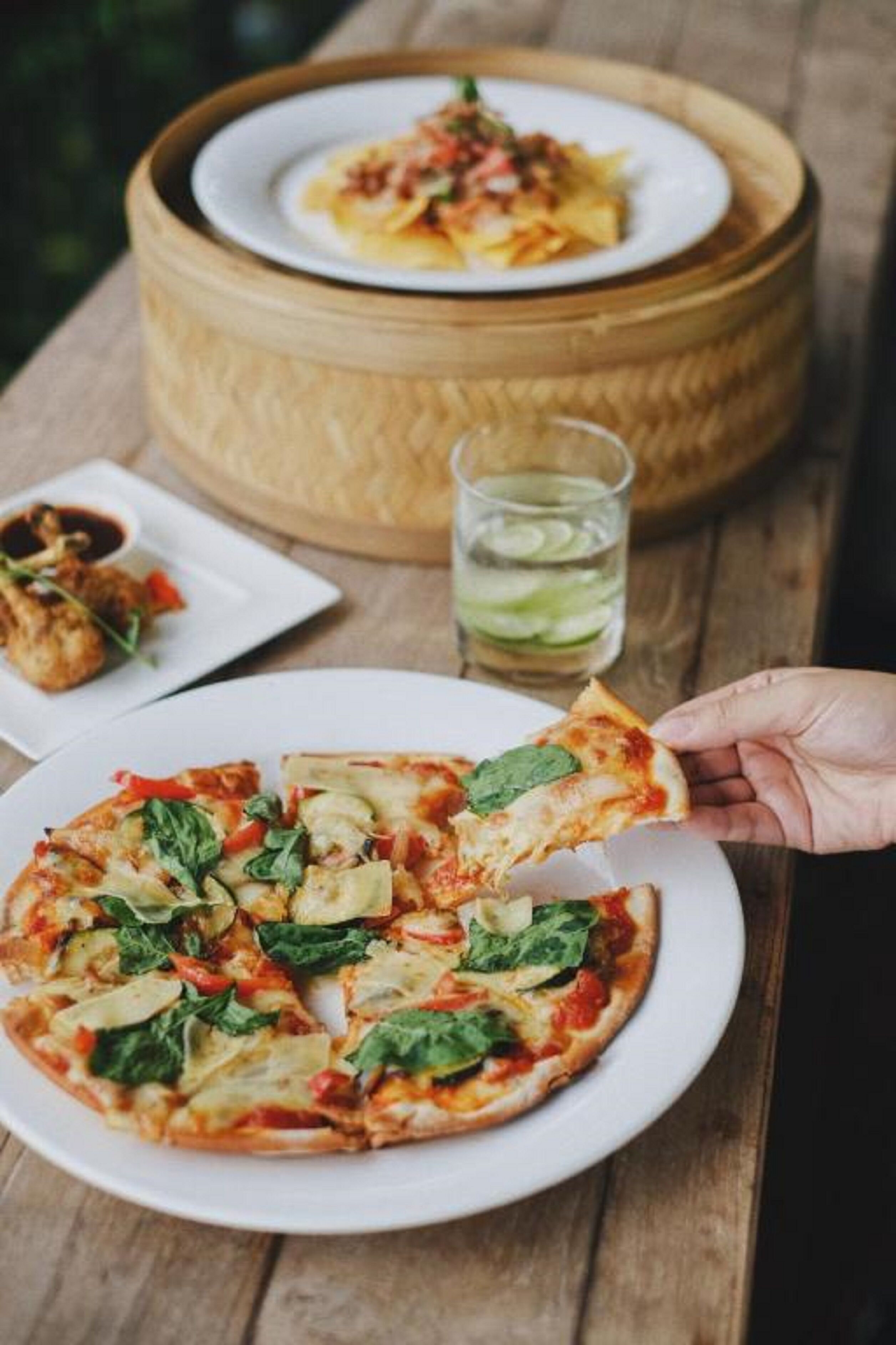 Pizza con espinacas y pimientos rojos sobre un plato blanco, una mano sosteniendo una porción, y un vaso de agua con rodajas de pepino sobre una mesa de madera.