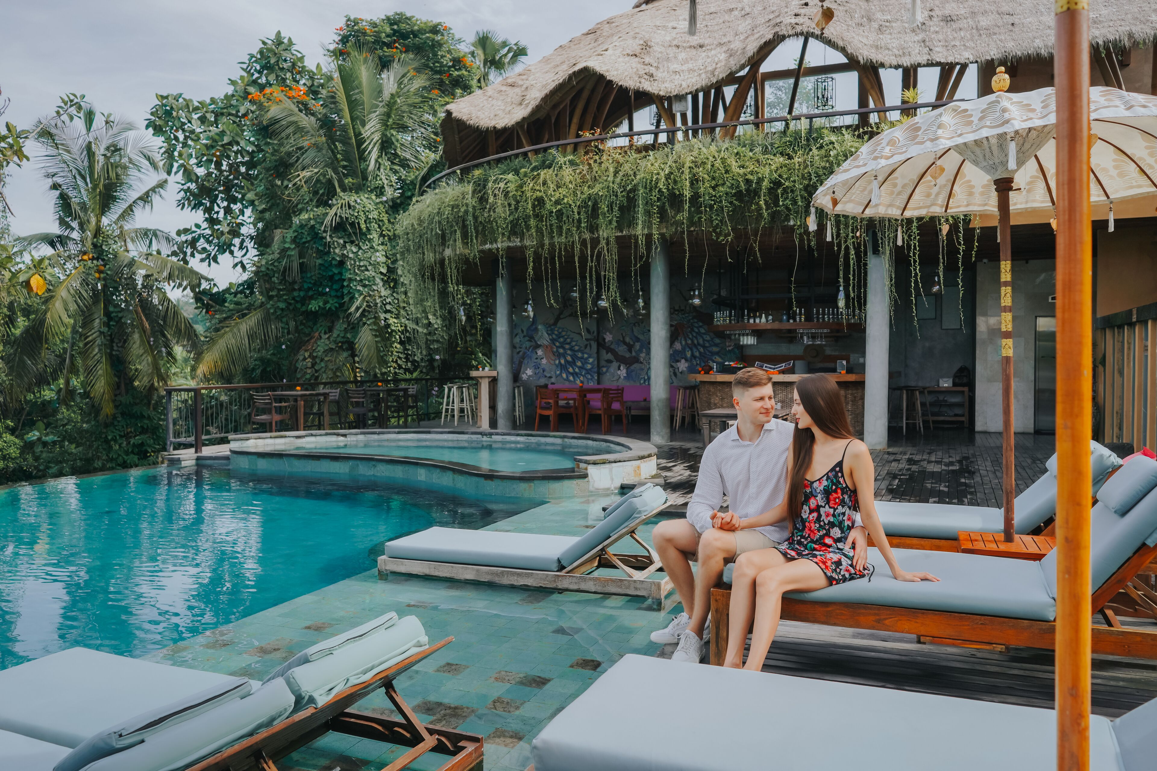 Un couple est assis sur des chaises longues près d’une piscine, sous un toit de chaume, avec des plantes suspendues.