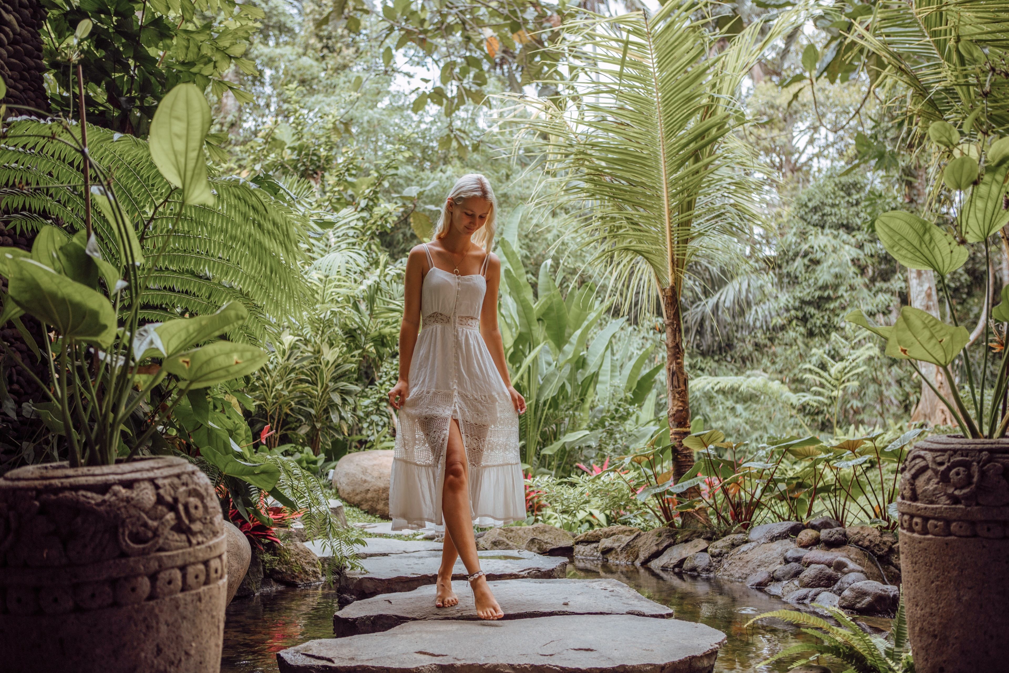 A woman in a white dress standing on a stone bridge in a lush garden with various plants and a small stream.