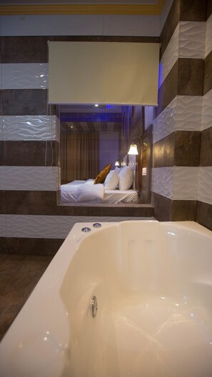 A modern bathroom with a large bathtub, striped walls, and a mirror reflecting a bedroom with a bed and pillows. - Hotel Kings Regency (Jwalamukhi)