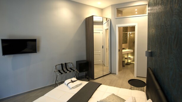 A modern hotel room with a bed, a TV, a seating area, a wardrobe, and a bathroom visible through an open door.
