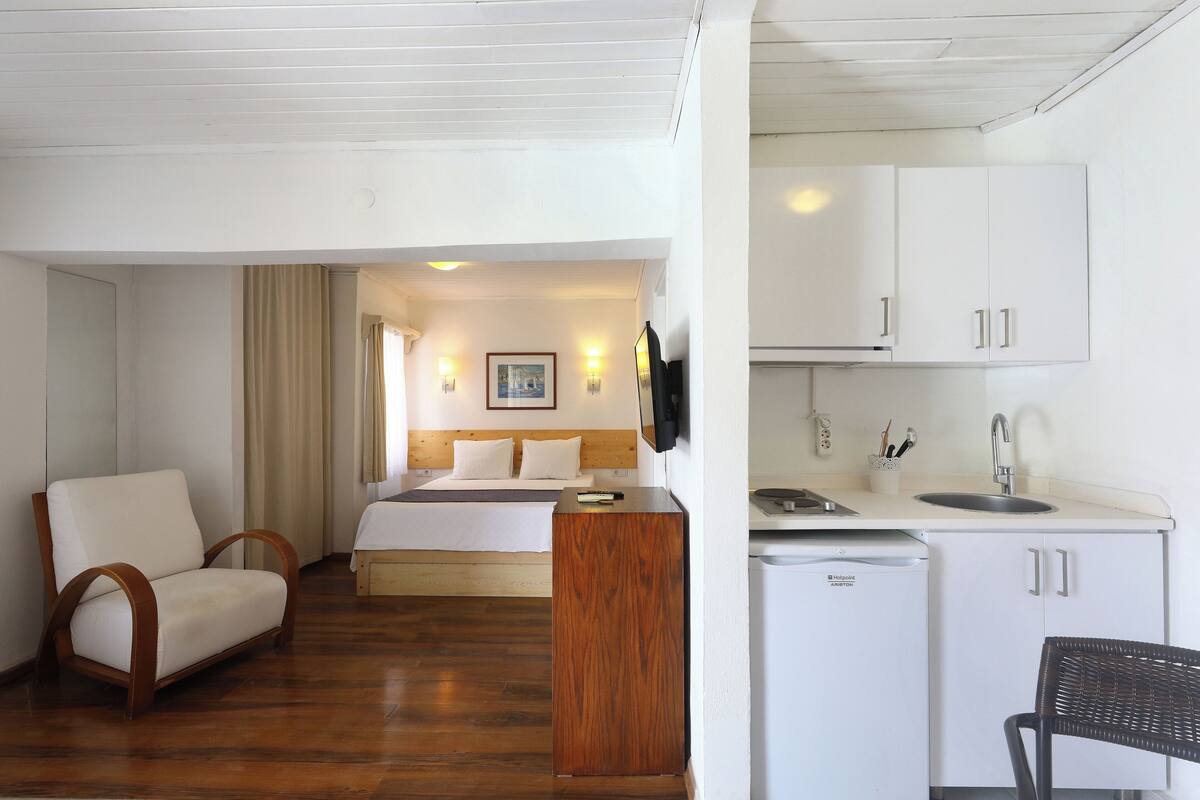 Una habitación de hotel con cama, una silla de madera, una pequeña kitchenette y un escritorio.