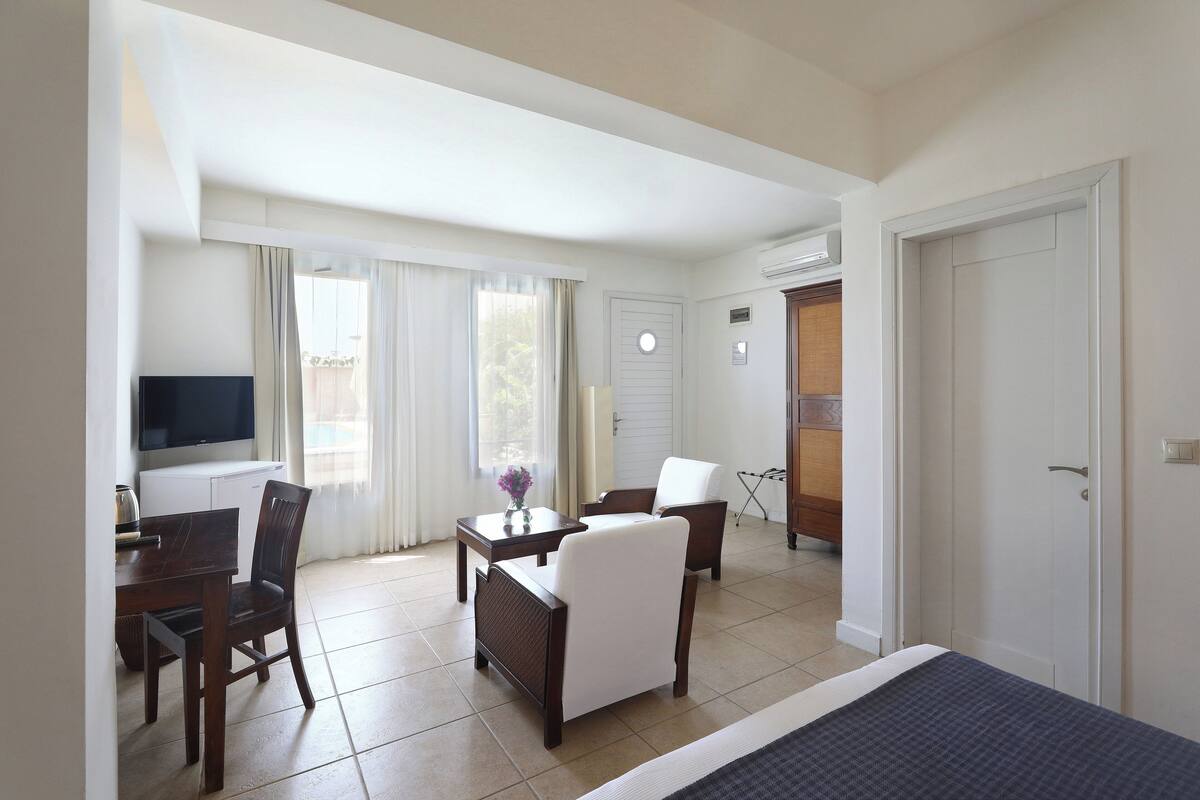 Habitación de hotel con cama, televisor, mesa de comedor con sillas y una mesita con un florero.