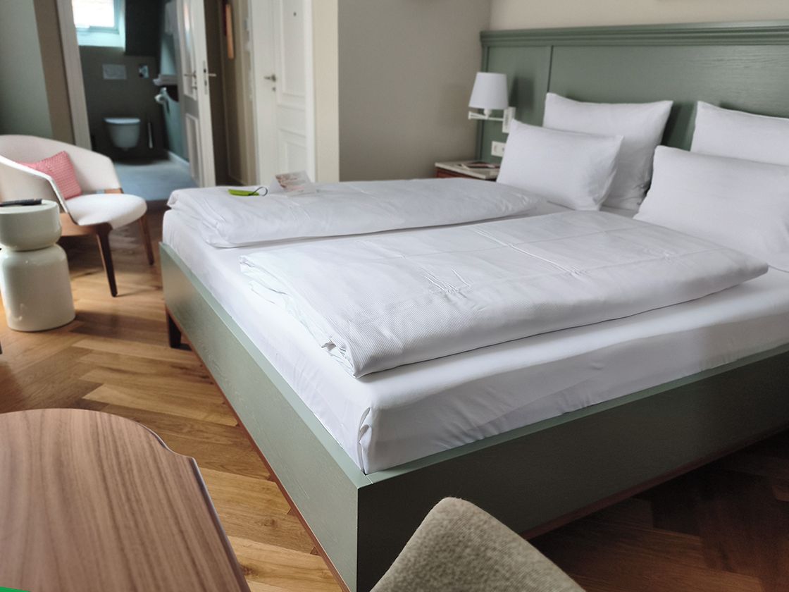 Una cama doble con sábanas blancas, una mesita de noche con lámpara y una silla con cojín.