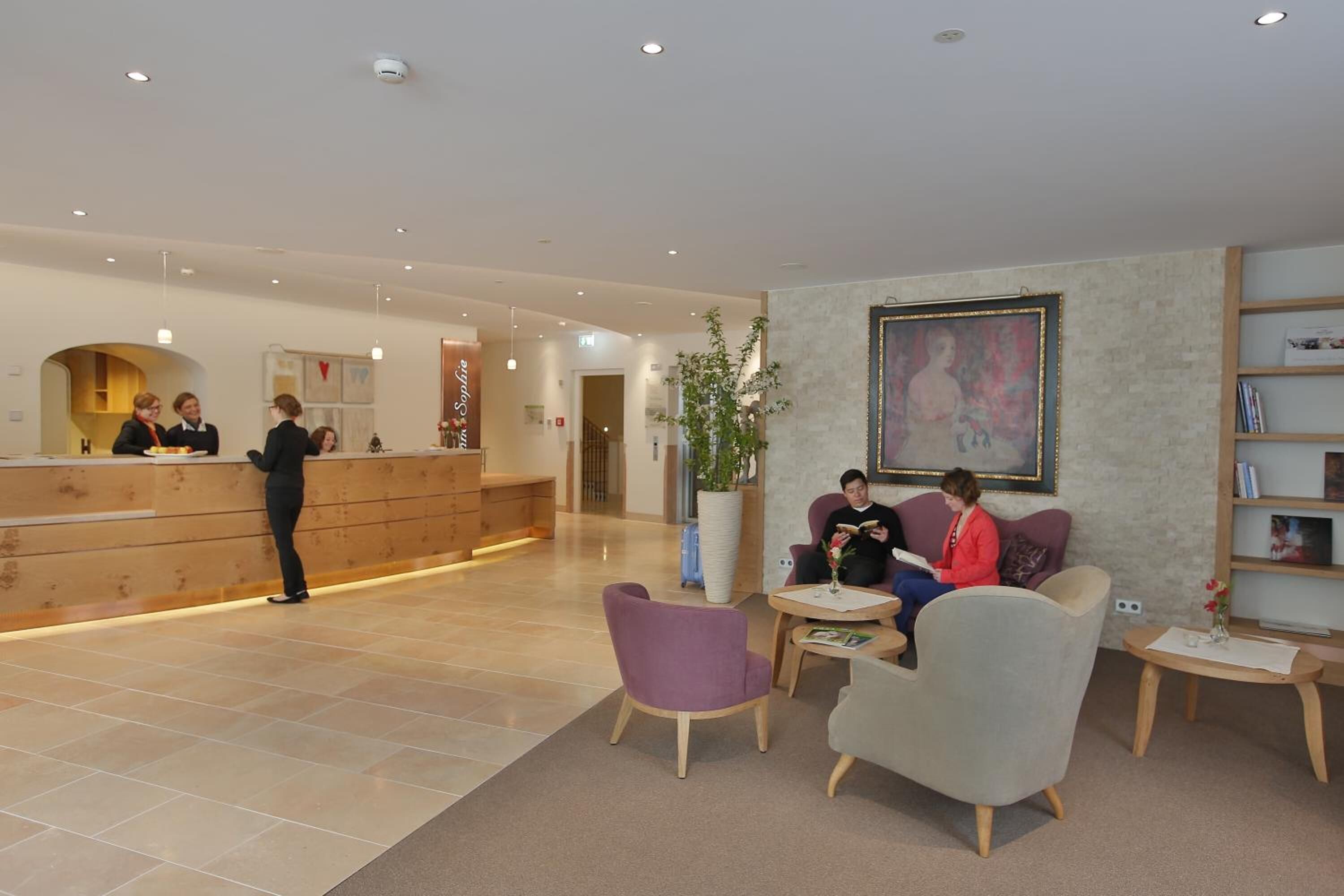A modern hotel lobby with a reception desk, seating area, and a large painting on the wall.