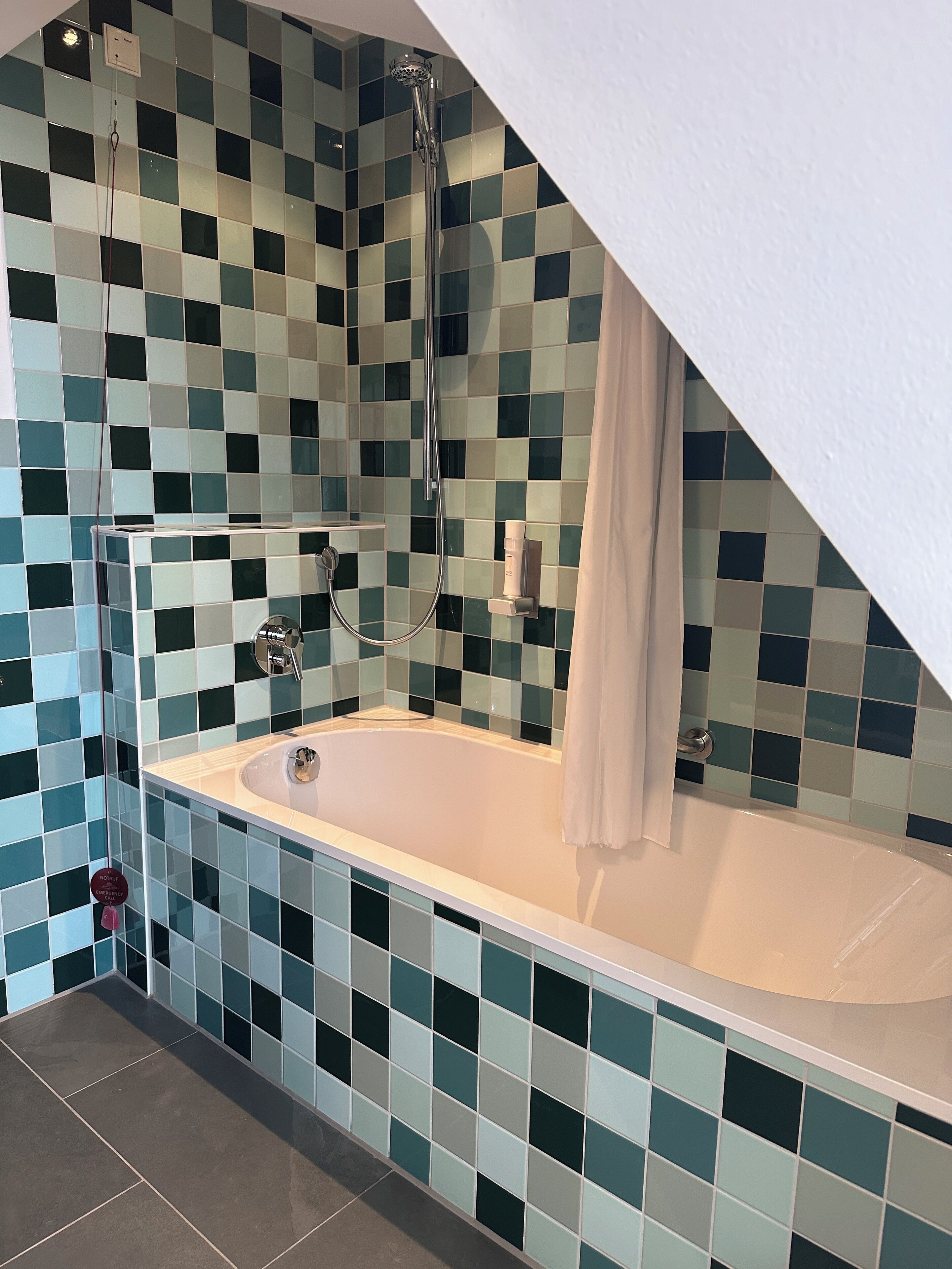 Un baño con bañera, paredes de azulejos, ducha y cortina blanca.