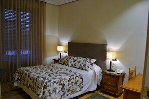 Hotelkamer met een bed, twee bedlampjes, een nachtkastje met telefoon, een houten stoel en een raam met jaloezieën.