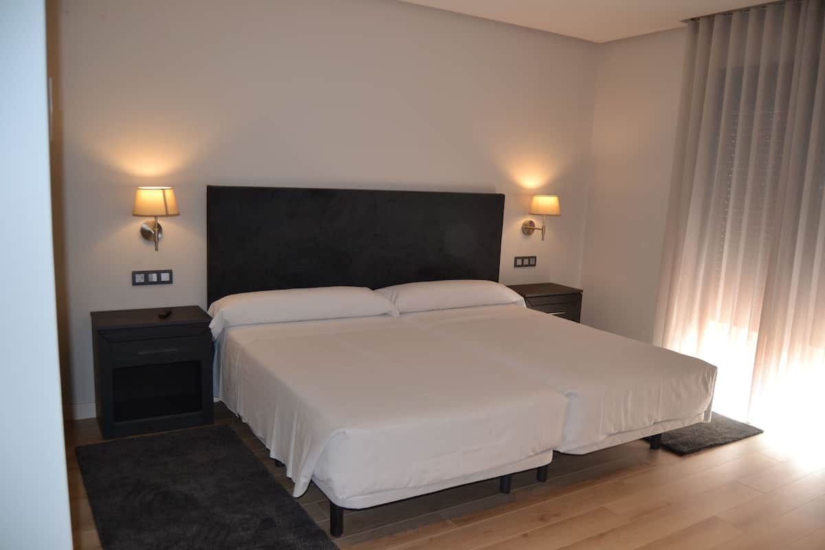 Una cama bien hecha con sábanas blancas, dos mesitas de noche con lámparas y un cabecero oscuro.