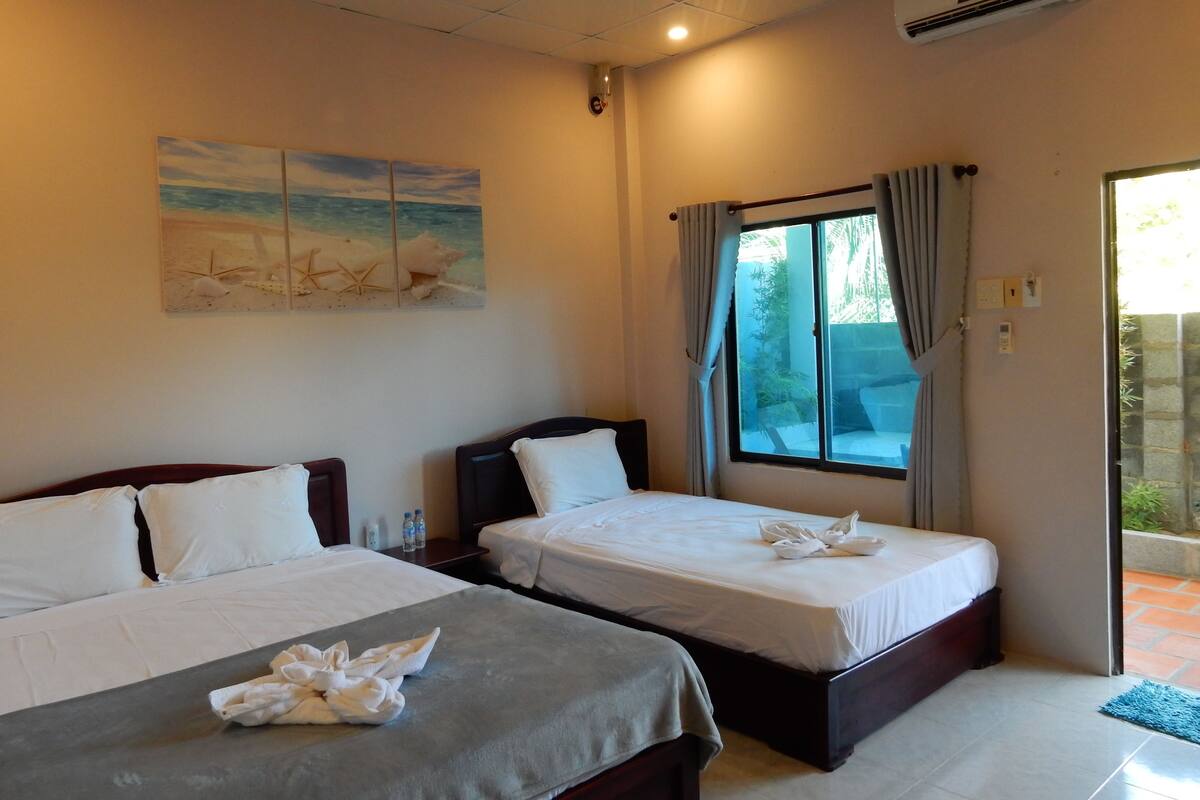Habitación de hotel con dos camas, un ventanal con cortinas, un cuadro con temática playera y un equipo de aire acondicionado.