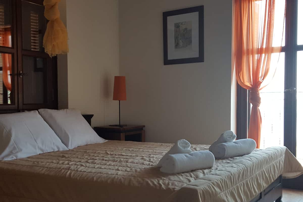 Una cama bien tendida con sábanas blancas y dos toallas enrolladas, una lámpara de noche y un cuadro en la pared.