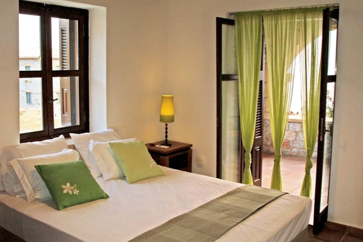 Un dormitorio con cama, almohadas, una mesita de noche con lámpara, una ventana con cortinas y una puerta abierta que da a un balcón.