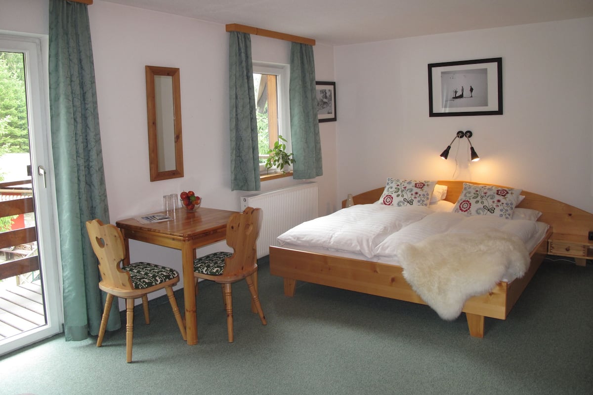 A bedroom with a wooden bed, two chairs, a table, a window with curtains, a radiator, and a framed picture on the wall.