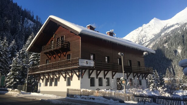 Una cabaña de madera con balcón, rodeada de árboles y montañas cubiertas de nieve.