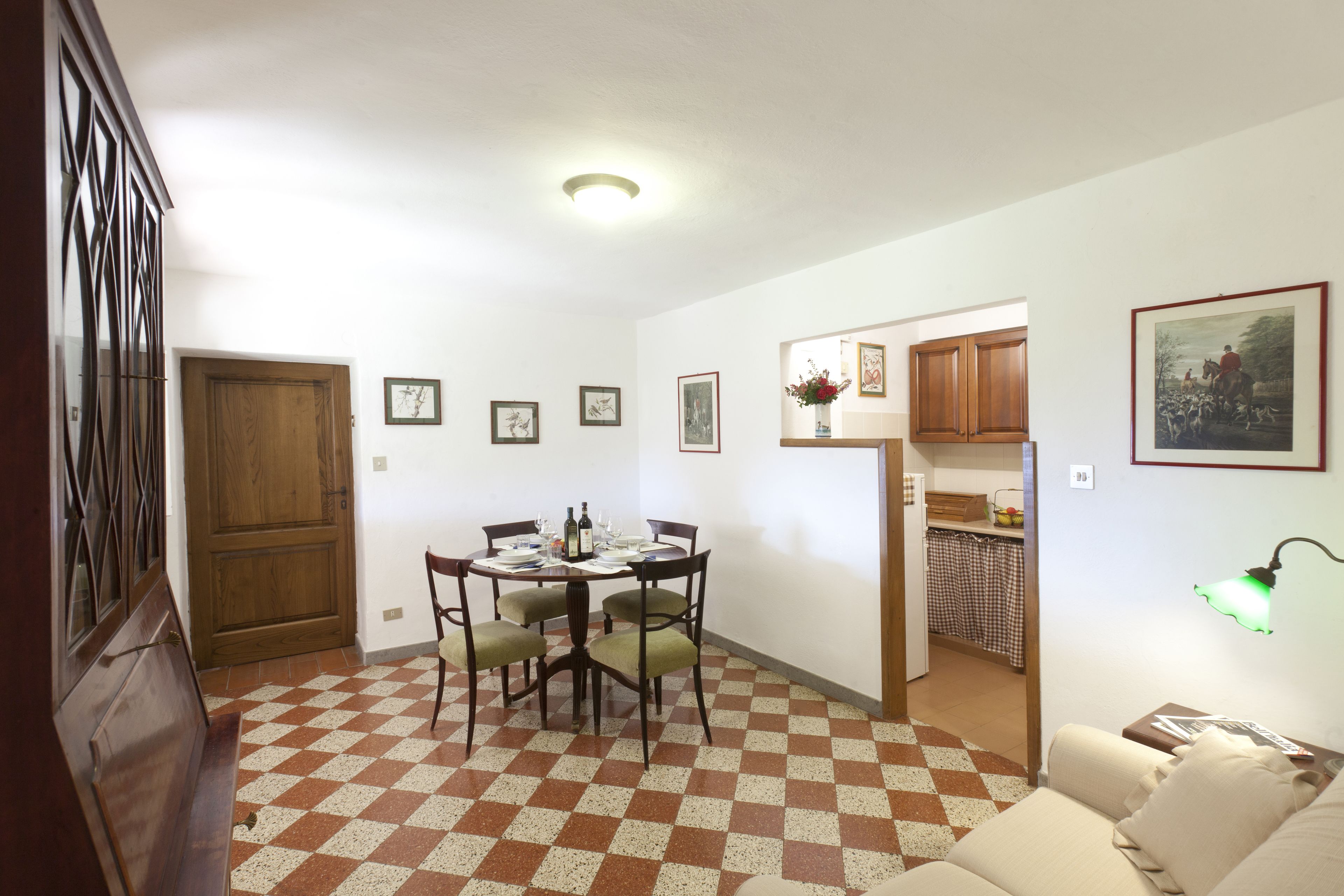 Een eetruimte met een zwart-witte vloer, een ronde tafel met stoelen, een houten deur en een keuken zichtbaar door een open doorgang.