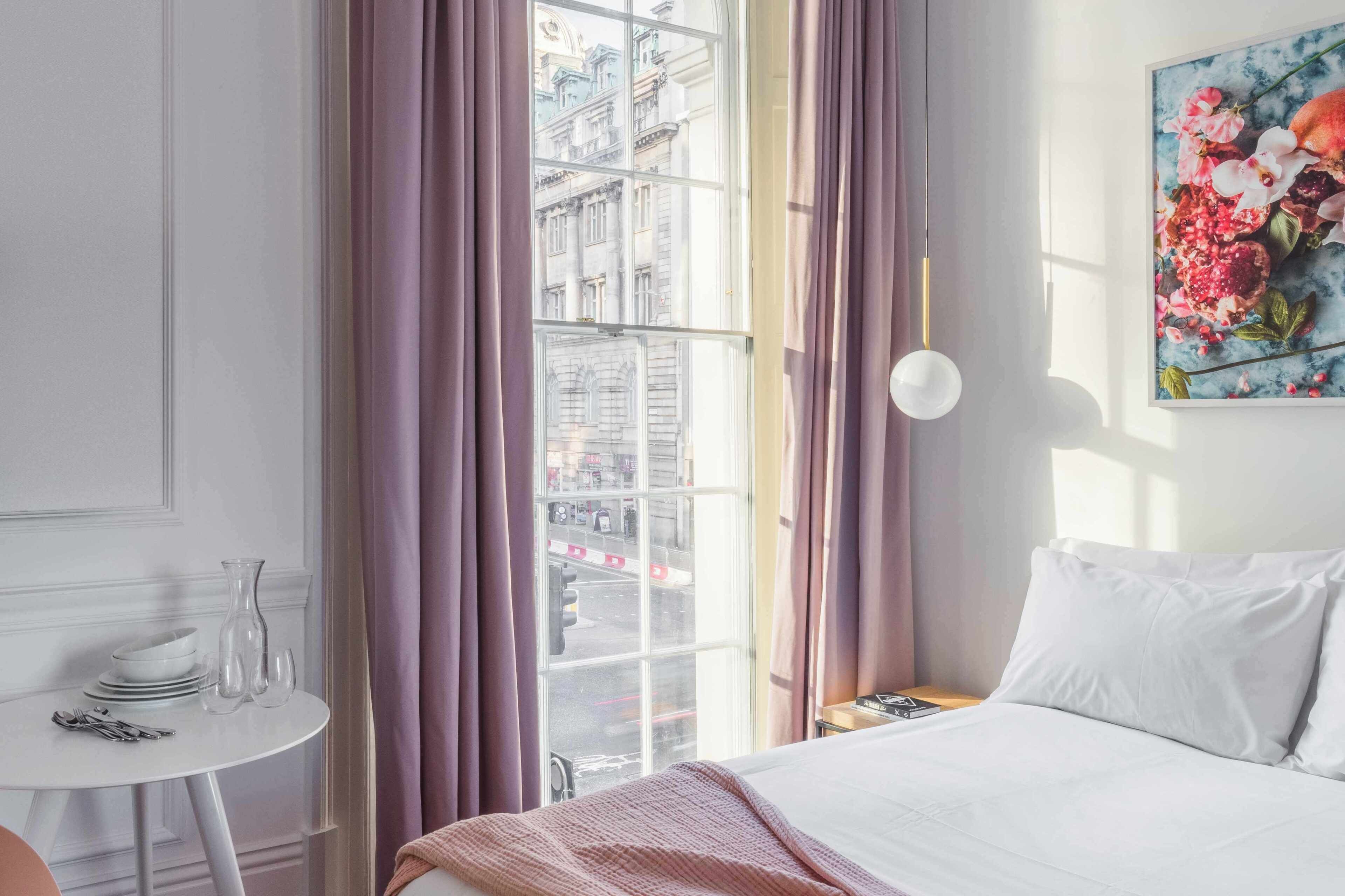 A hotel room with a bed, a small table with a vase, a window with curtains, and a painting on the wall.