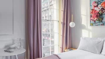 A hotel room with a bed, a small table with a vase, a window with curtains, and a painting on the wall.