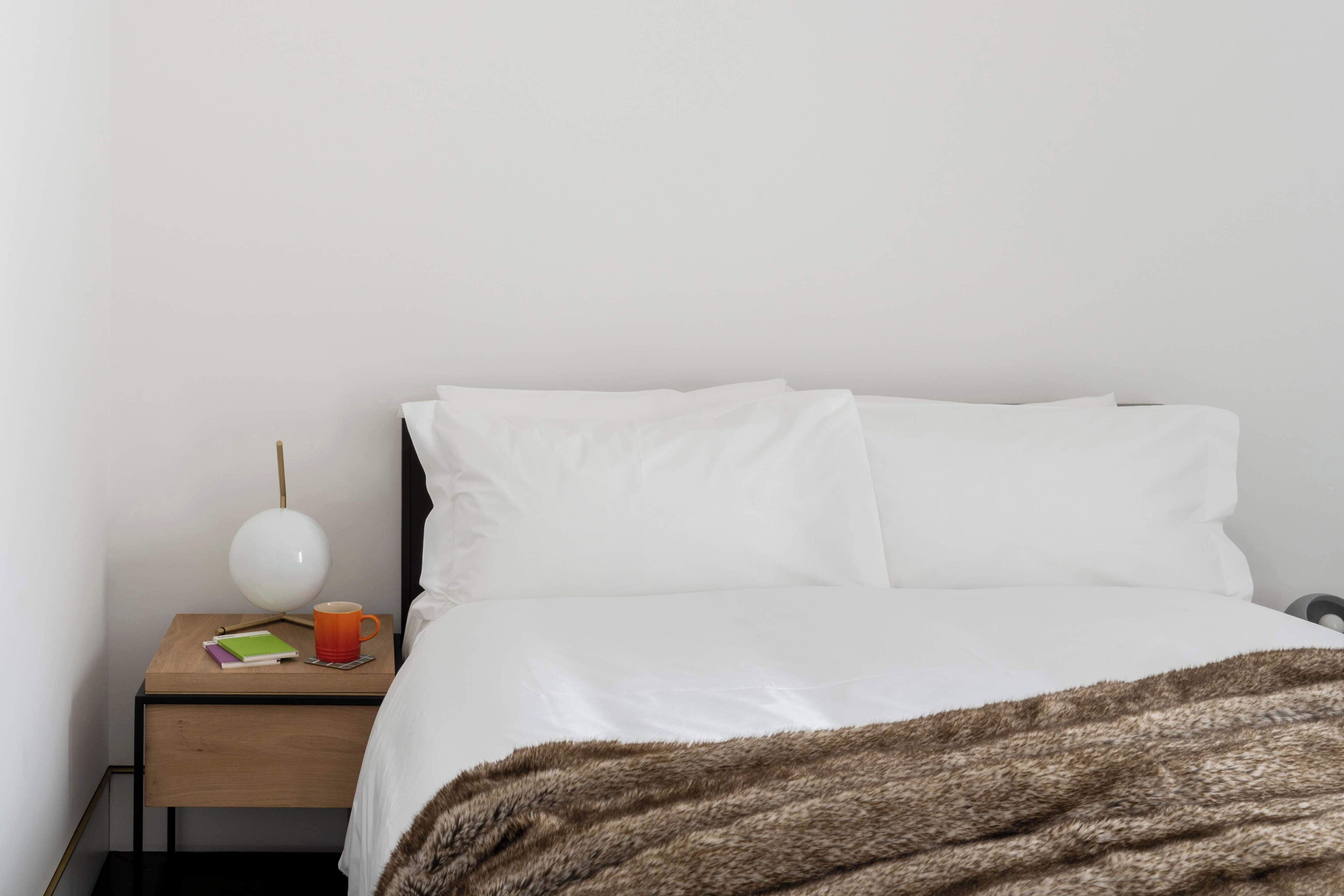 A simple, clean hotel room with a bed, a bedside table, a lamp, and a small table with a book and a mug.
