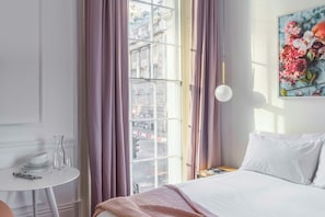 A hotel room with a bed, a small table with a vase, a window with curtains, and a painting on the wall.