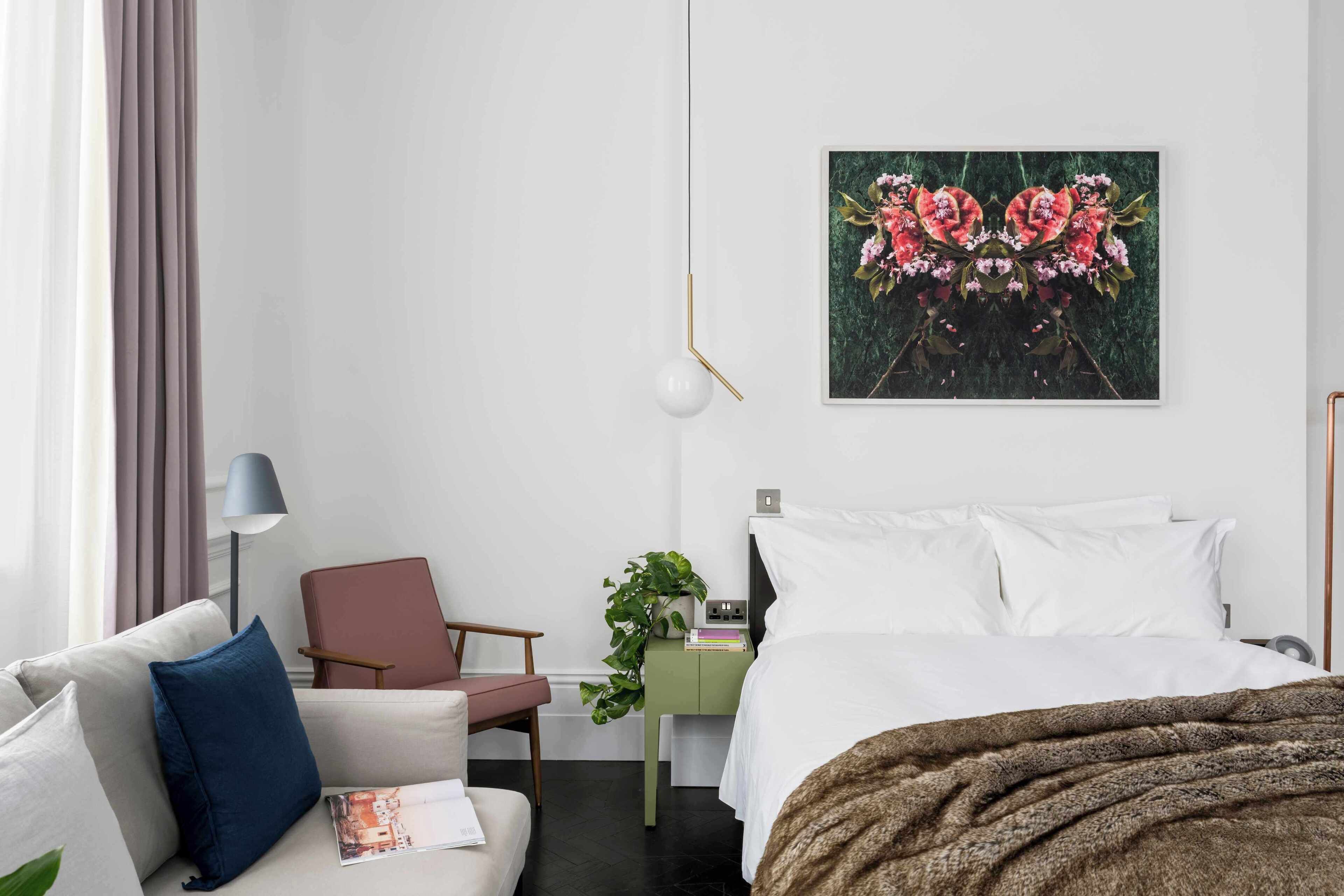 A modern hotel room with a bed, a sofa, a chair, a desk with a plant, and a wall-mounted artwork.