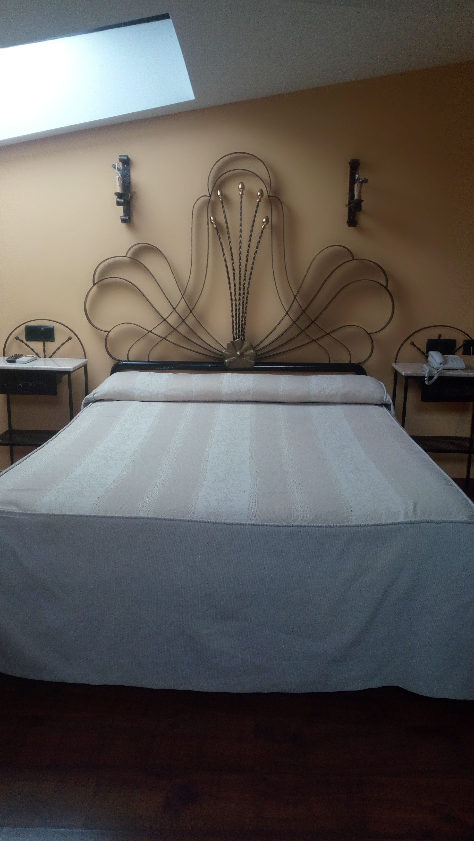 Cama com cabeceira adornada com motivos florais, duas mesinhas de cabeceira e um claraboia acima.