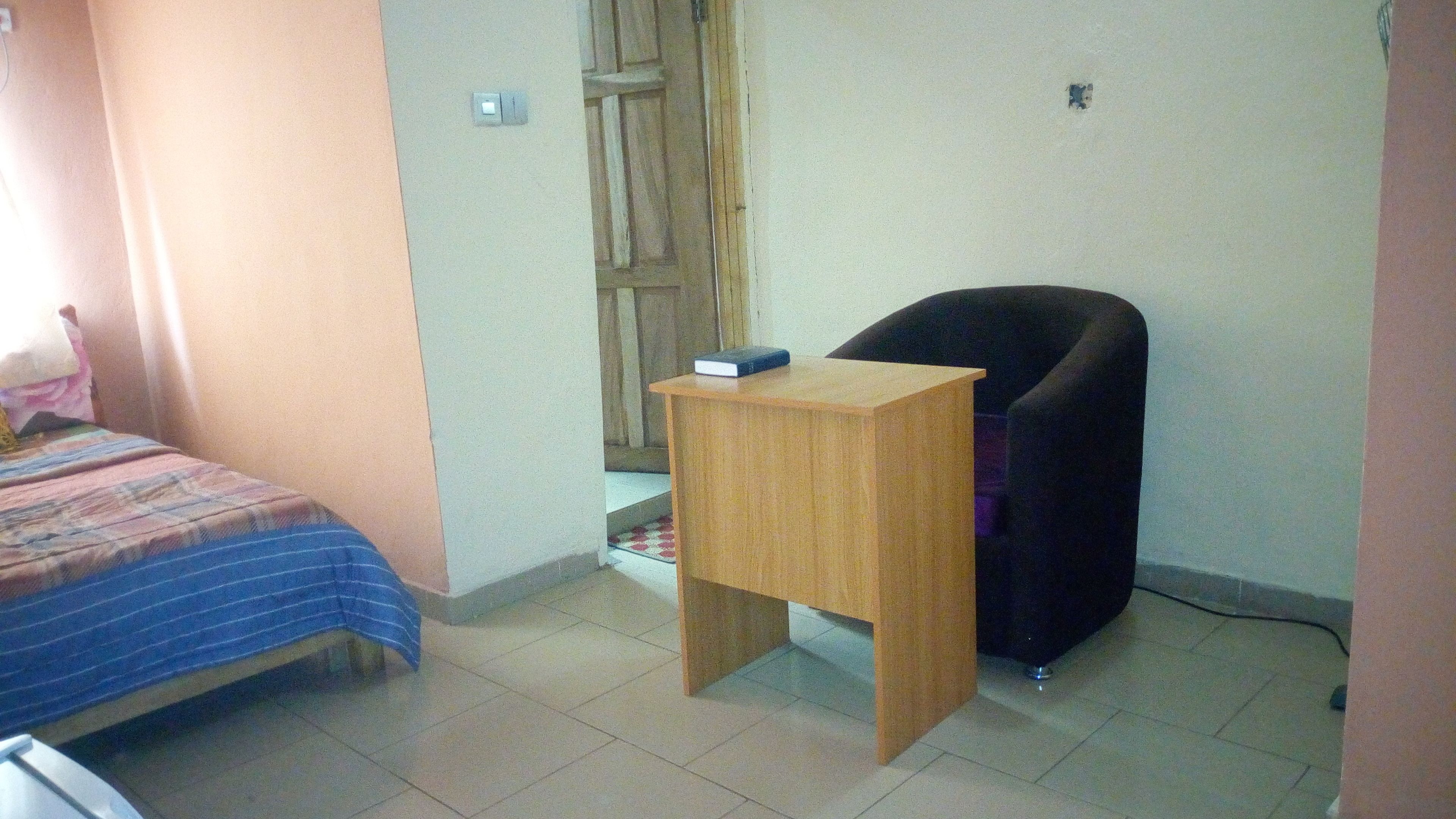 A small, sparsely furnished room with a single bed, a wooden nightstand, a black office chair, and a door leading to another room.