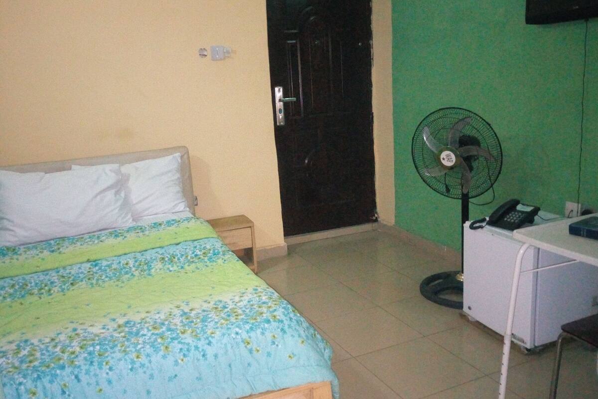 A room with a bed, a desk, a chair, a fan, and a door.