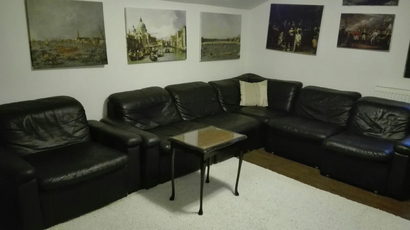 A living room with a black leather sectional sofa, a glass coffee table, and four paintings on the wall.