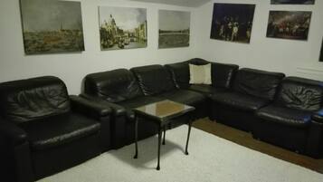 A living room with a black leather sectional sofa, a glass coffee table, and four paintings on the wall.
