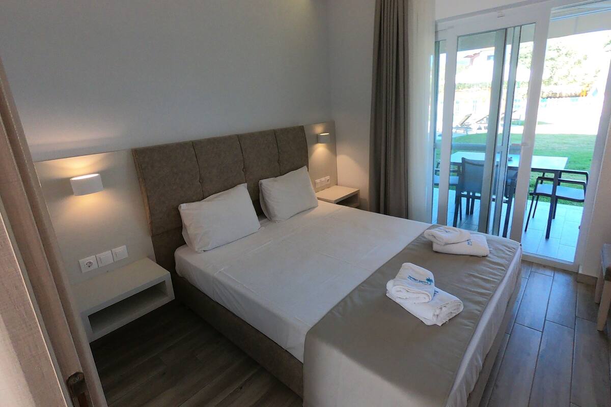Ein Hotelzimmer mit einem großen Bett, einem Nachttisch, einer Fenstersturz und Blick ins Freie durch eine Glastür.