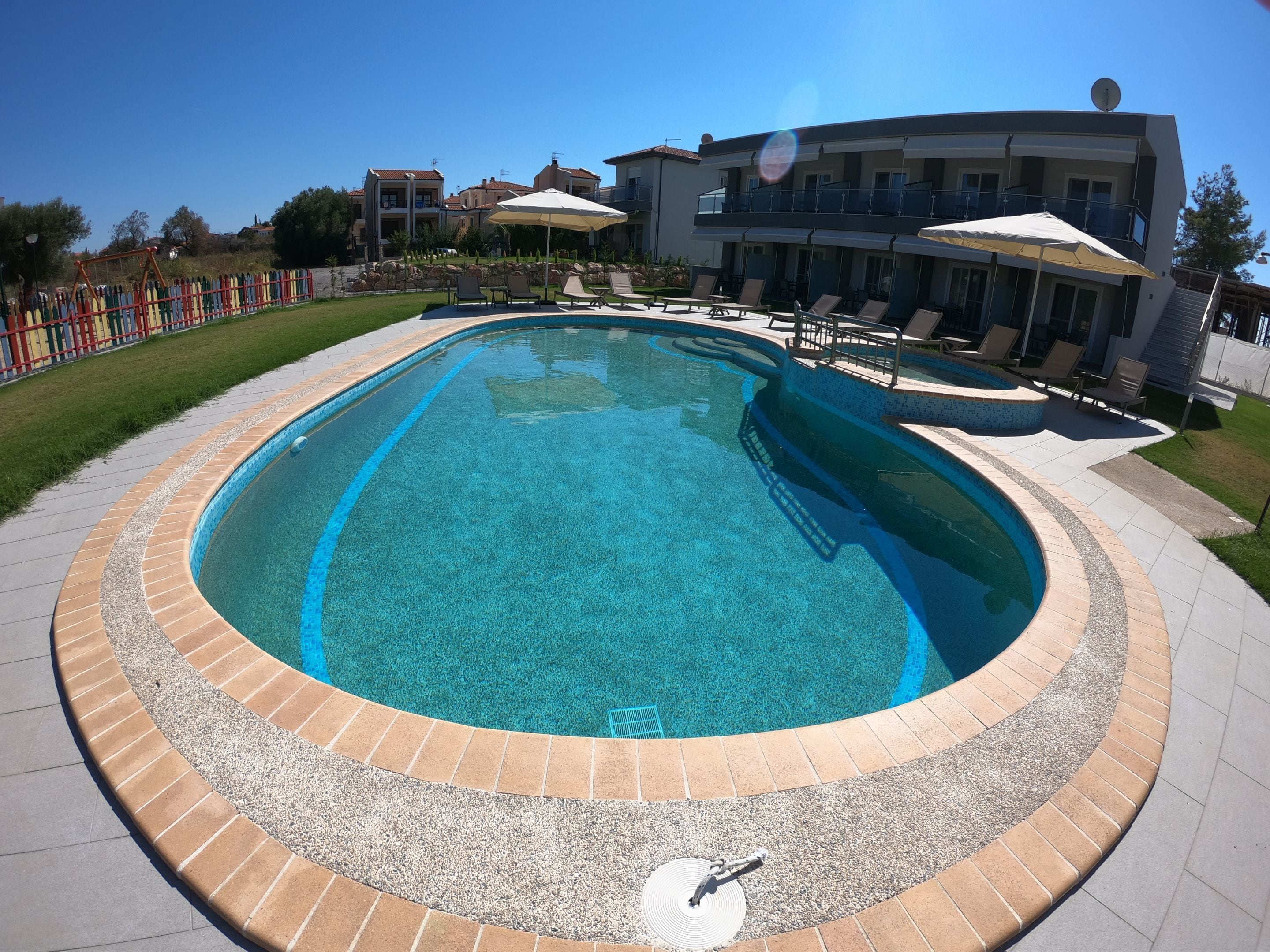 A swimming pool with a tiled edge, surrounded by a brick border. There are lounge chairs and umbrellas near the pool.