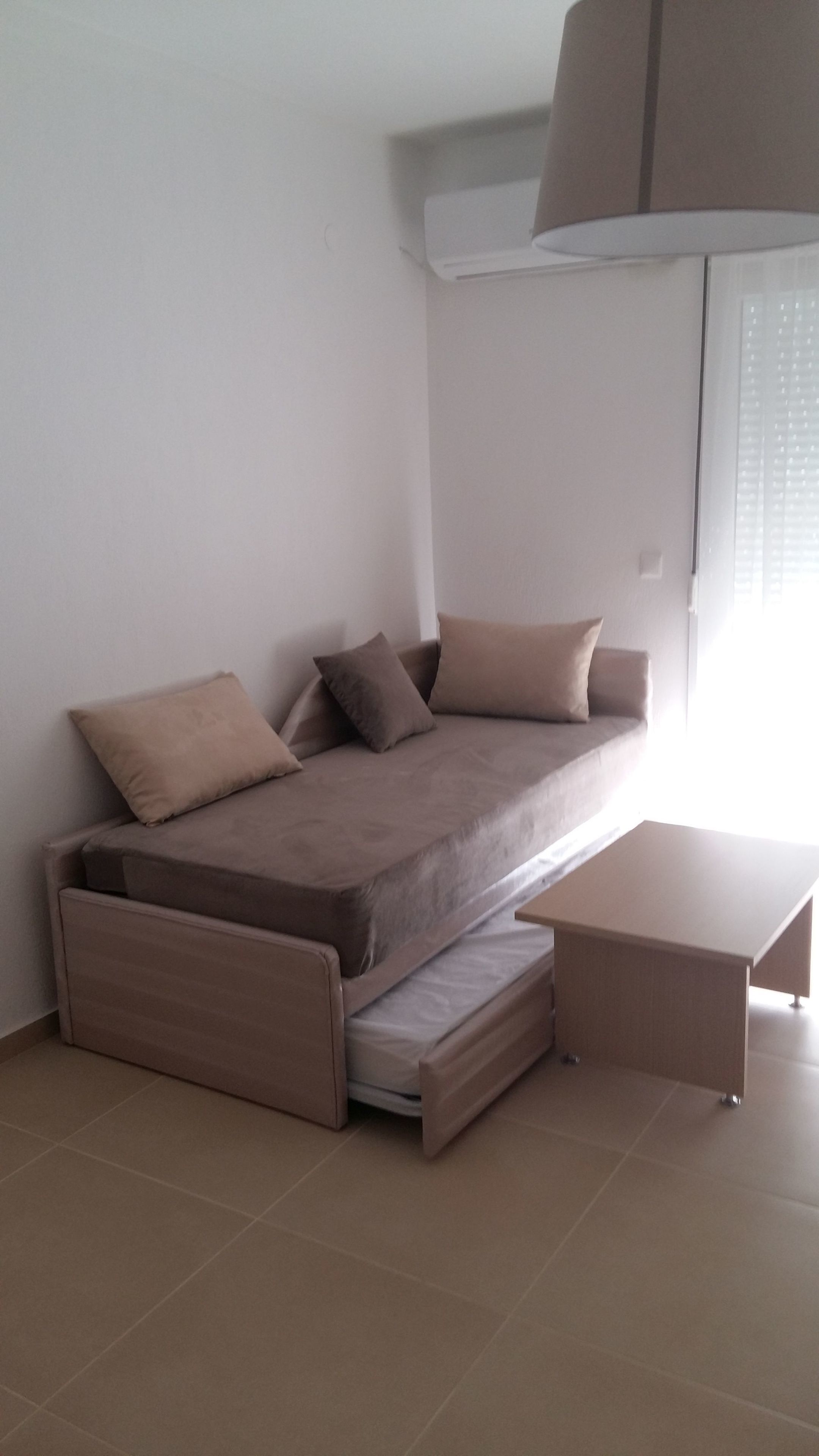 A compact, modern room with a convertible sofa bed, a small table, and a window with blinds.