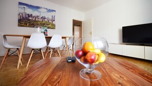 Een eethoek met een houten tafel, een glazen schaal met fruit, witte stoelen, een schilderij van een stadsgezicht en een televisie op een witte kast.
