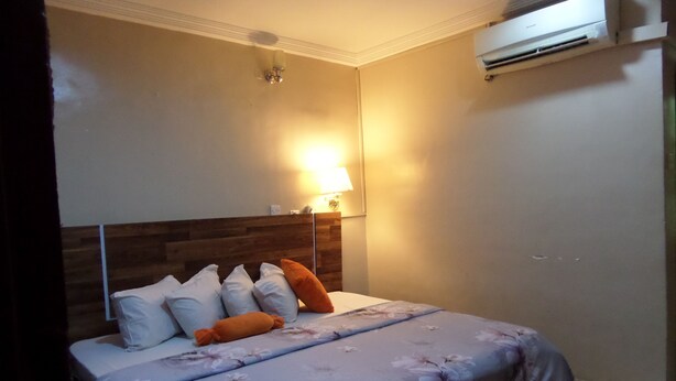 A bedroom with a bed, pillows, a nightstand, a wall-mounted air conditioner, and a bedside lamp. - Koltol Guest House (Ibadan)