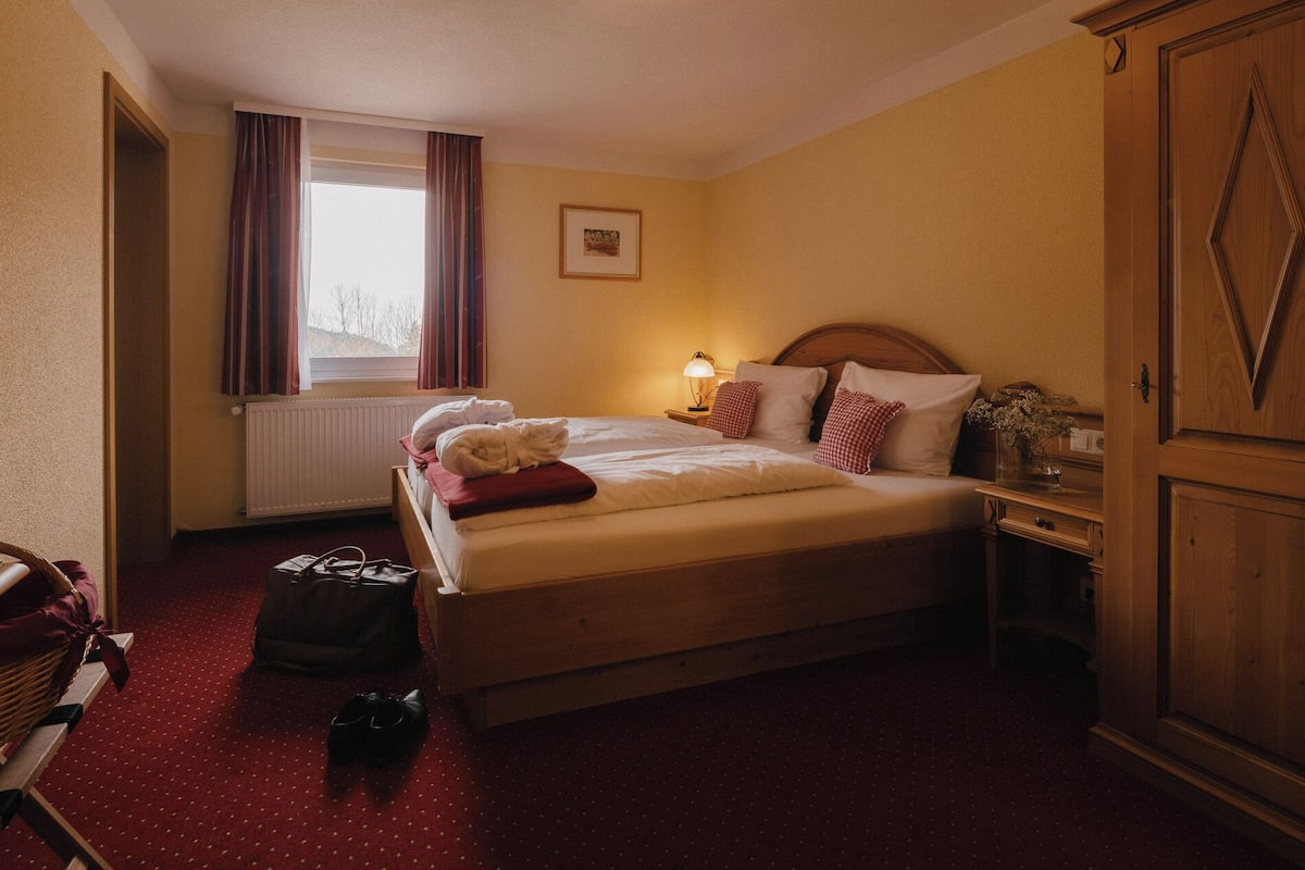 Quarto de hotel com cama, mesa de cabeceira, abajur, radiador, janela com cortinas, guarda-roupa de madeira e um quadro na parede.