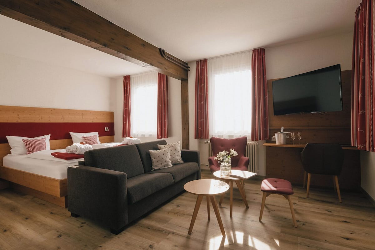 Quarto de hotel com uma cama, um sofá, uma mesinha, uma cadeira, uma televisão e um radiador.