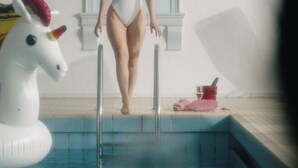 Una mujer con traje de baño blanco y sombrero negro sale de una piscina con un flotador de unicornio y toallas rosas cerca.