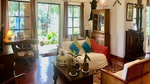 Una sala de estar acogedora con un sofá, sillones, una mesita de centro y un aparador. Hay ventanas con cortinas, una lámpara y adornos en las paredes.