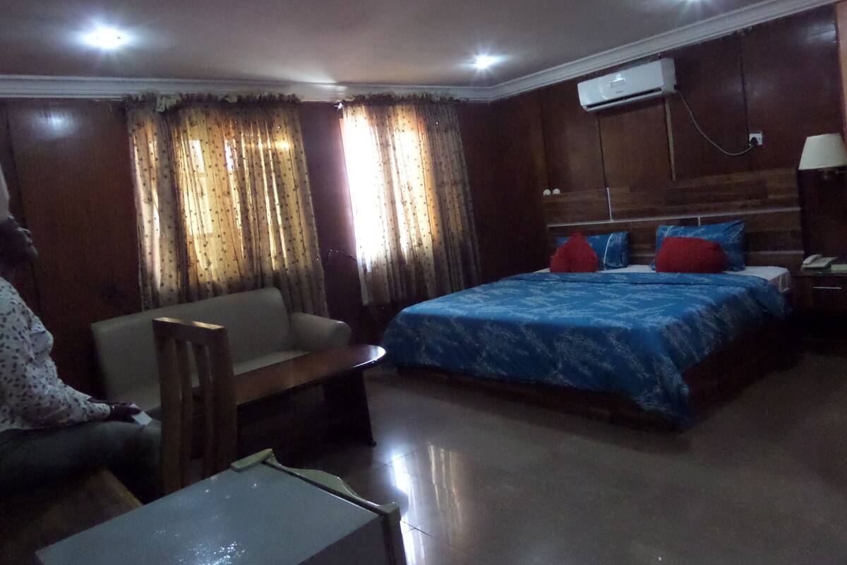 A hotel room with a bed, a sofa, a small table, and an air conditioning unit on the wall.