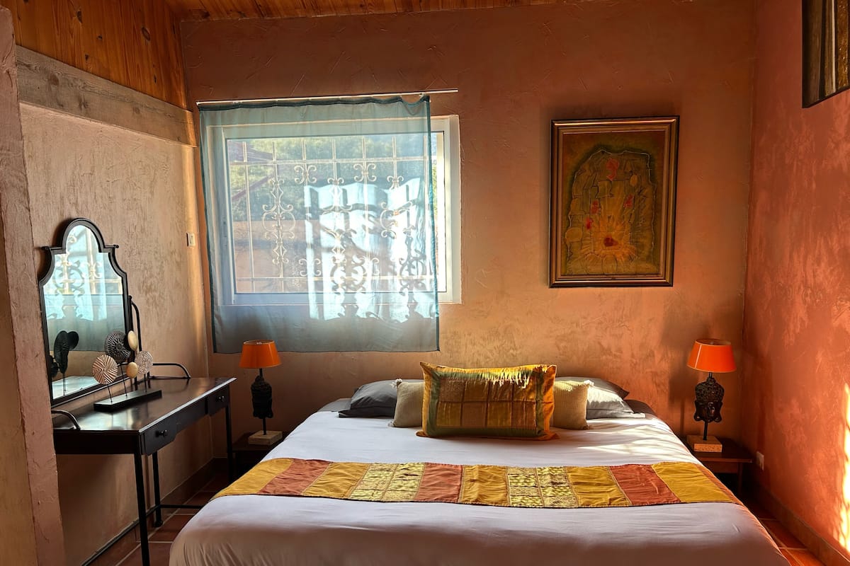 A bedroom with a bed, two bedside lamps, a window with decorative iron bars, a mirror, and a framed artwork on the wall.