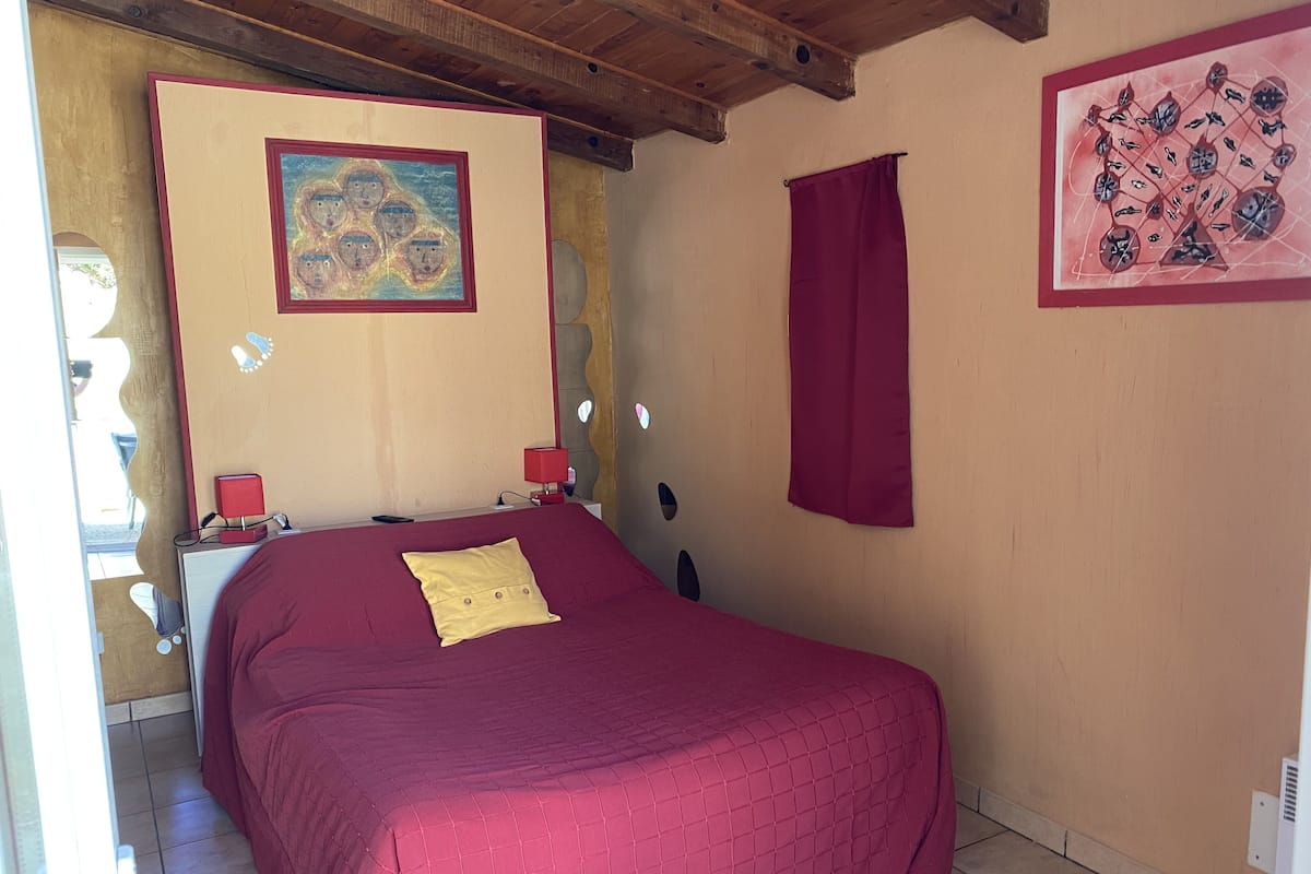 A bedroom with a bed, a nightstand, a red lamp, and a wall painting.