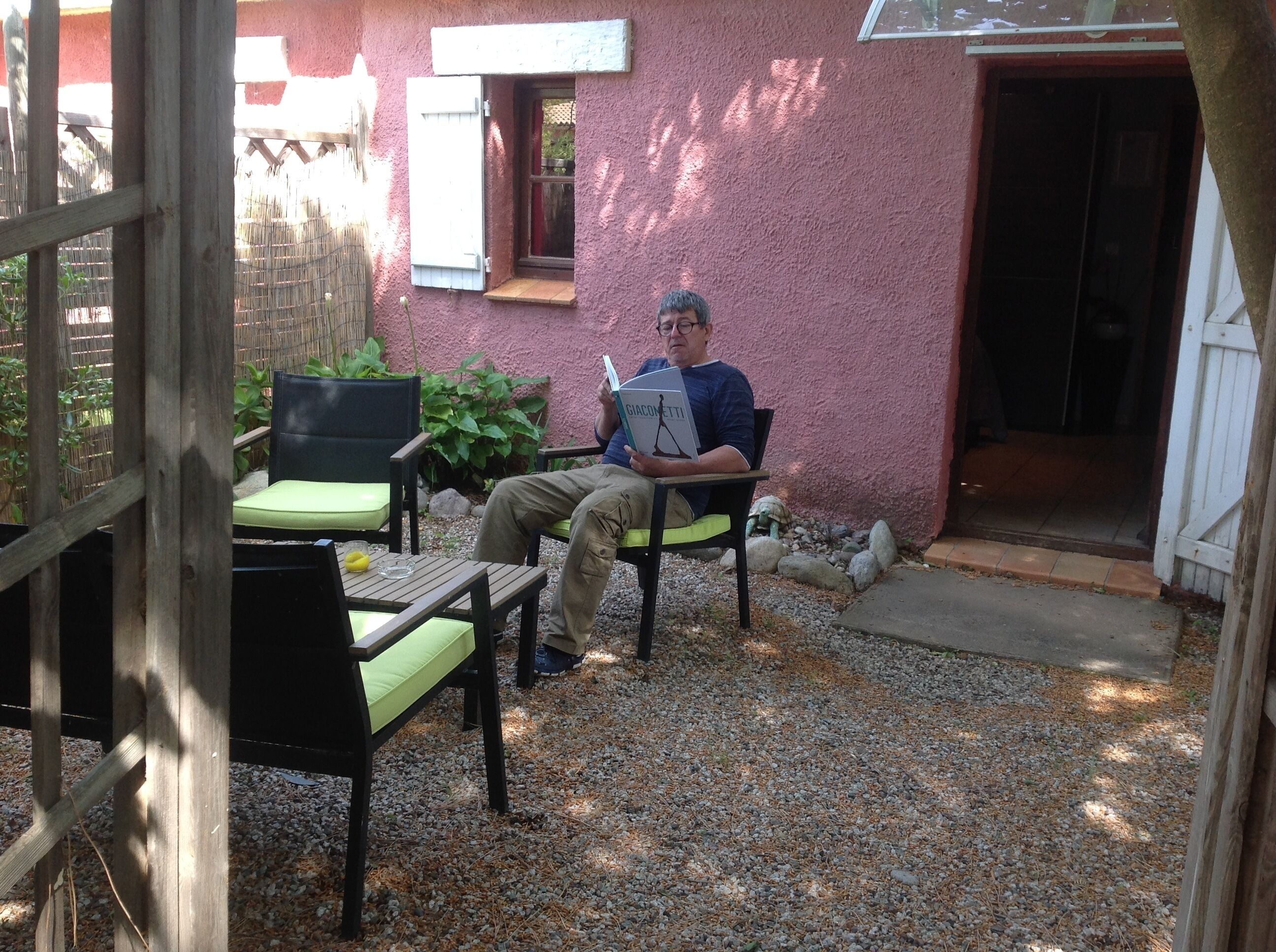 Ein Mann sitzt auf einem Stuhl und liest ein Buch auf einer Terrasse mit Holztisch, Stühlen und einem kleinen Garten.
