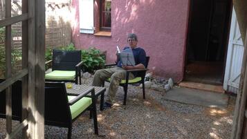 Ein Mann sitzt auf einem Stuhl und liest ein Buch auf einer Terrasse mit Holztisch, Stühlen und einem kleinen Garten.