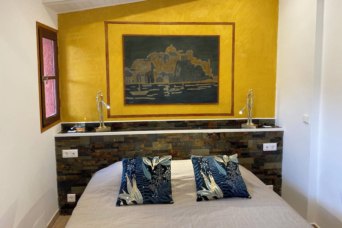 A hotel room with a bed, a painting on the wall, and a stone wall behind the bed.