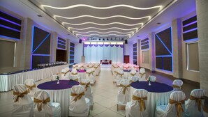A banquet hall with round tables and chairs, a stage with a backdrop, and modern lighting fixtures. - Elite Palazzo (Aluva)