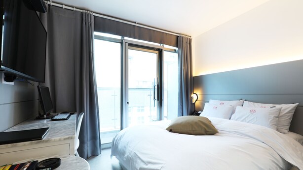 A hotel room with a bed, a desk with a computer, a TV, and a window with curtains. - Hotel yaja wirye (Seongnam)