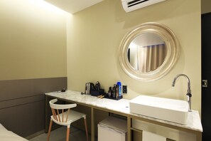 Casa de banho de hotel compacta com lavatório, espelho e uma pequena mesa com produtos de higiene pessoal.