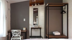 A modern hotel room with a bed, a desk with a chair, a bathroom with a sink and mirror, and a wooden shelf with towels. - Lemon tree Studio Stellenbosch Central (Stellenbosch)