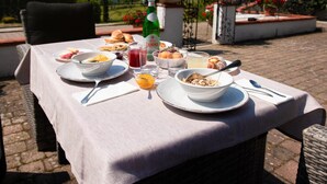 An outdoor dining table set with breakfast items, a bottle of wine, and a scenic view of a garden and mountains.